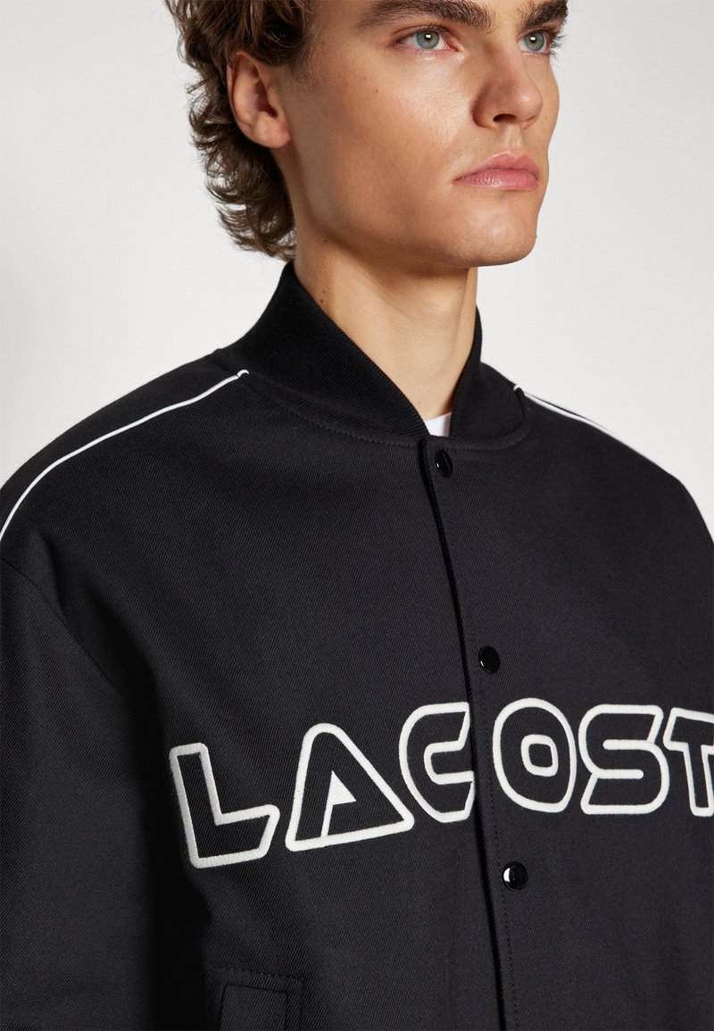 Veste noire avec col montant, présentant le lettrage brodé "LACOSTE" en blanc et des fermetures à boutons, fabriquée en tissu texturé.