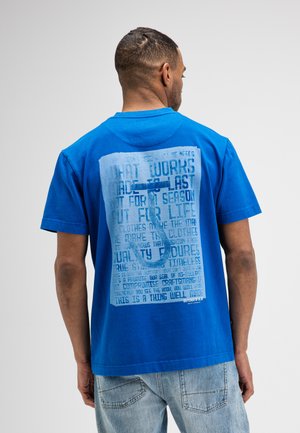 Man draagt een blauwe T-shirt met grote bedrukte tekst en grafisch ontwerp op de rug, gecombineerd met lichtblauwe spijkerbroek.