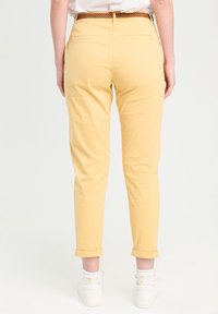 Pantalon en coton jaune avec une coupe fuselée, ourlets retroussés et poches arrière, assorti à des baskets blanches et des chaussettes à motifs.