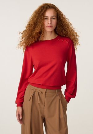 Vrouw met lang krullend rood haar, die een rode trui en hoge bruine broek met een geknoopt riem draagt, staande met één hand in de zak.