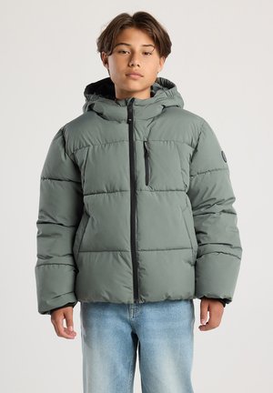 Teenager trägt eine grüne gepolsterte Jacke mit Kapuze und schwarzem Reißverschluss, steht vor einem schlichten hellen Hintergrund.