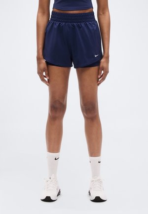 Personne portant un short de sport bleu marine, des chaussettes blanches avec des logos noirs et des baskets blanches, debout sur un fond blanc.
