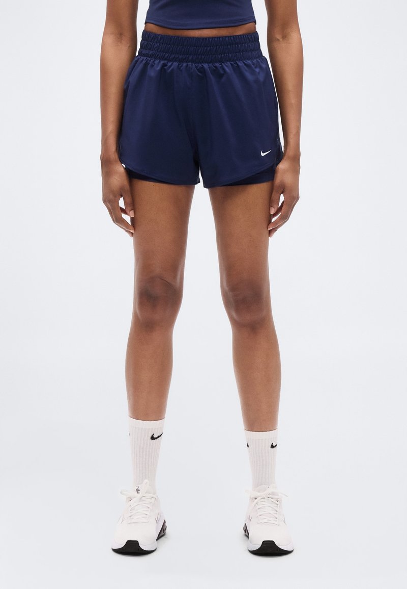 Personne portant un short de sport bleu marine, des chaussettes blanches avec des logos noirs et des baskets blanches, debout sur un fond blanc.