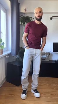 Mladik s platinasto plavom kosom i bradom, odjeven u bordo majicu, bijele hlače i crno-bijele cipele, stoji u modernoj sobi.