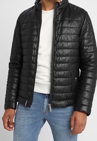 Man i svart vadderad skinnjacka över en vit skjorta och blå jeans, med händerna avslappnat vid sidorna.