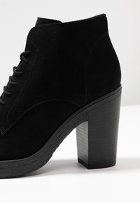 Botín de ante negro con punta redonda, diseño con cordones y tacón grueso negro. Presenta detalles texturizados cerca de la base.