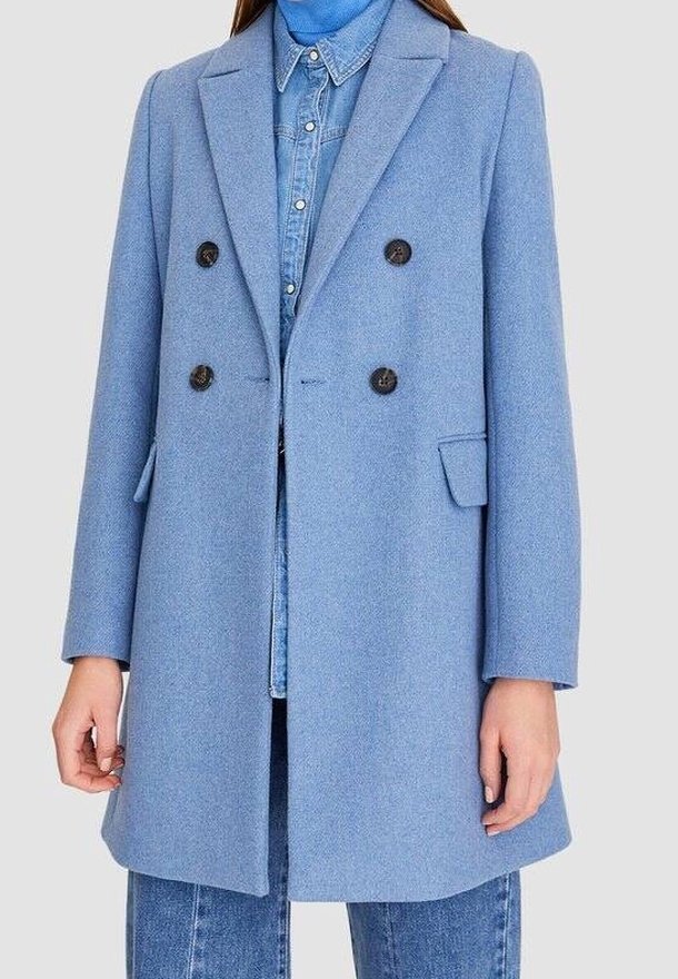 Hellblauer Wollmantel im doppelreihigen Design mit schwarzen Knöpfen, Reverskragen und seitlichen Taschen, über einem Denimhemd getragen.