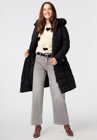 Zwarte gewatteerde jas met een met bont gevoerde capuchon, beige sweater met hartpatronen, grijze wijde broek en laarzen met luipaardprint.