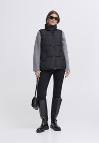 Zwarte gewatteerde bodywarmer met een hoge kraag en grijze gebreide mouwen. Draagt over zwarte broek en gecombineerd met zwarte laarzen tot de knie en een zonnebril.