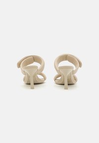 Beige høyhælte sandaler med en glatt tekstur, med to overlappende stropper og en slank, buet hæl. Minimalistisk design og åpen bak.