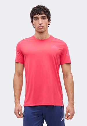 Hombre con cabello rizado que lleva una camiseta deportiva de manga corta de color rosa brillante y pantalones cortos azul oscuro, de pie frente a un fondo liso.