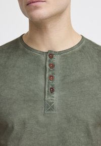 Grøn bomulds henley skjorte med rund halsudskæring, der har fem brune knapper og synlige sømdetaljer. Blød tekstur med et falmet udseende.