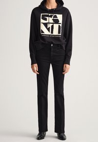 Sudadera negra con capucha y logo gráfico blanco, combinada con jeans negros de pierna recta y botas negras hasta el tobillo, sobre un fondo neutro.