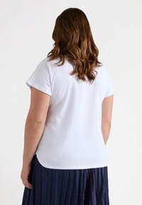 Camiseta blanca de manga corta con cuello redondo. Textura suave y dobladillo ligeramente curvado. La vista posterior muestra la longitud de los hombros y de la camiseta.