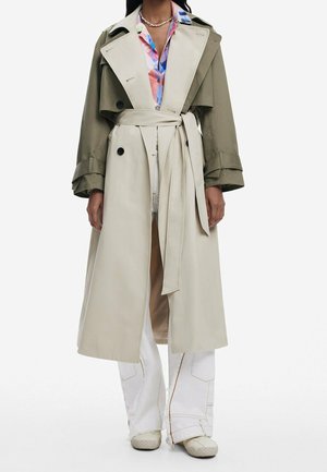 Persona con un abrigo trench bicolor con mangas verde oliva y cuerpo beige, atado a la cintura, pantalones blancos, camisa colorida y zapatos beige.