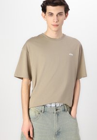 Tricou oversized khaki din bumbac moale, cu guler rotund și mâneci scurte, având un mic logo alb în zona stângă a pieptului.