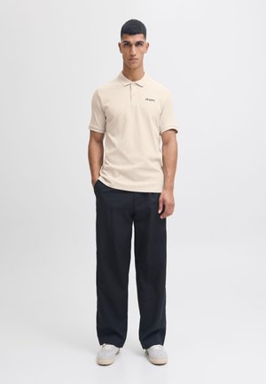 Beige-paitainen polo-paita lyhyillä hihoilla ja tekstuurilla, yhdistettynä löysiin mustiin housuihin ja harmaisiin tennareihin.