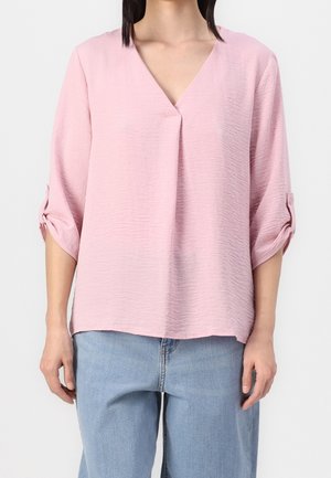 Femme portant un chemisier texturé rose clair à col en V avec des manches mi-longues retroussées et un jean en denim bleu clair sur un fond uni.