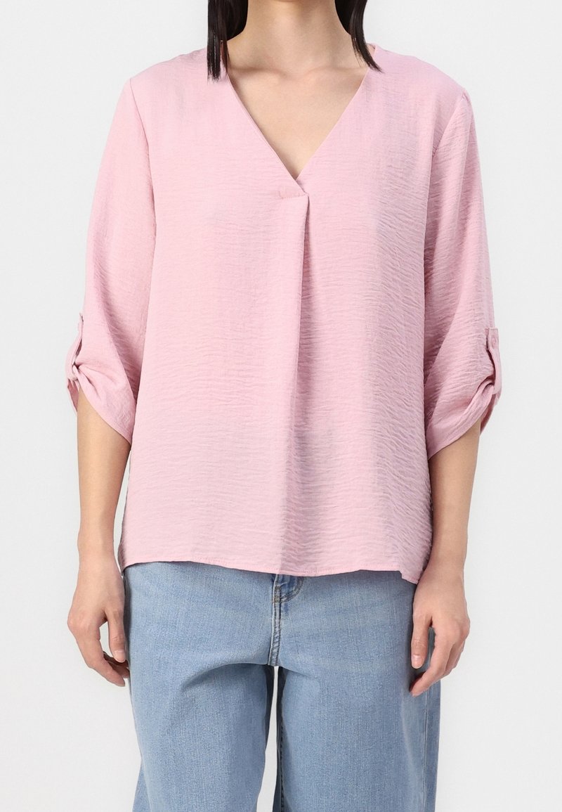 Femme portant un chemisier texturé rose clair à col en V avec des manches mi-longues retroussées et un jean en denim bleu clair sur un fond uni.