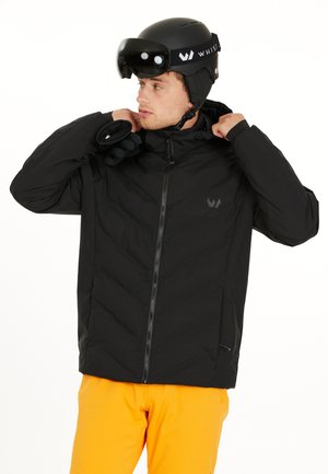 Schwarze isolierte Jacke mit Reißverschluss, ausgestattet mit einem hohen Kragen und einem Chevron-Design, kombiniert mit leuchtend orangen Hosen und einem schwarzen Helm.