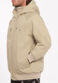 Beige hættetrøje med justerbare snoretræk, ribbede manchetter og sidelommer, med en glat, mat overflade og diskret logo-detalje.