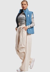 Plavi podstavljeni prsluk, majica dugih rukava s printom, kremaste široke hlače i bijele tenisice. Istaknuti dizajnerski elementi uključuju logo na prsluku i džepove.