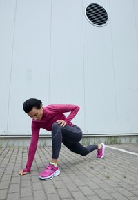 Vêtements de sport comprenant un haut ajusté à manches longues rose et un legging gris foncé avec des détails perforés. Chaussures de course roses vives avec des semelles blanches.