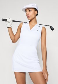 Bílý skládaný golfový šaty s V-výstřihem, s malým logem na hrudi a přiléhavým střihem, doplněné o bílou čepici a rukavici.