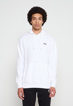 Joven con pelo rizado que lleva una sudadera con capucha blanca de FILA y pantalones deportivos negros de FILA, de pie frente a un fondo claro y liso.