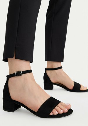 Pies con sandalias de tacón cuadrado y tiras de tobillo negras combinadas con pantalones negros recortados con aberturas laterales sobre un fondo claro.