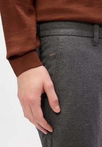 Grå uldragsbukser med en glat tekstur, der har en subtil orange syning. Hånden hviler på taljen, ingen synlig hardware.