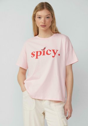 Pink cotton t-shirt featuring a red "spicy." text with a chili pepper accent. Loose fit and short sleeves, paired with light-colored pants.