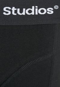 Schwarze Stretchstoff-Boxershorts mit einem breiten elastischen Bund, der das Wort "studios" in weißen Buchstaben zeigt. Mit flachen Nähten und einer glatten Textur.