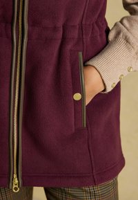 Fleece-Weste in tiefem Burgunderrot mit einem goldenen Reißverschluss, seitlichen Taschen und grauen Besätzen. Gerippte beigefarbene Sweater-Ärmel sind sichtbar.