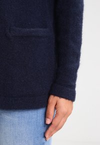 Navyblauer Wollcardigan mit strukturiertem Finish, langen Ärmeln und einer vorderen Aufsetztasche. Nahaufnahme des Ärmels und des Taschenbereichs.