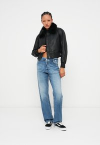 Casaco bomber de couro preto com colarinho de pelagem, design curto, combinado com calças de ganga de perna reta em azul claro e sapatilhas pretas com detalhes brancos.