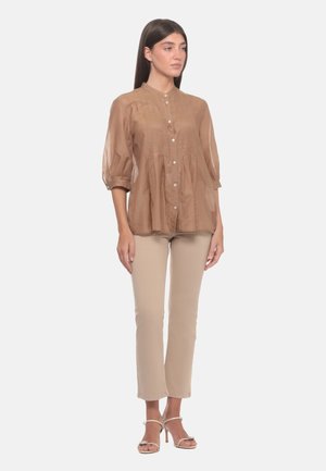 Donna in piedi che indossa una camicetta leggera marrone chiaro trasparente con bottoni, pantaloni beige aderenti e sandali beige con tacco alto su sfondo semplice.