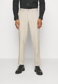 Pantaloni beige con vestibilità dritta, realizzati in tessuto morbido, dotati di una piastra frontale e passanti per cintura, abbinati a scarpe in pelle nere.