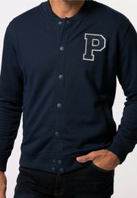 Homme portant une veste boutonnée bleu marine avec un grand écusson texturé en forme de "P" sur la poitrine gauche, la main glissée dans la poche d'un jean sombre.