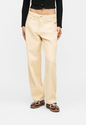 Persoon draagt losse beige broek, donker cropped top met lange mouwen en bruine sneakers met witte veters, staand tegen een effen achtergrond.