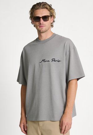 Man met schildpadzonnebril, grijs oversized T-shirt met tekst "Mira Paris" en beige broek, staand tegen een effen witte achtergrond.