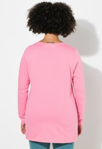 Pitkähihainen tunika kirkkaan vaaleanpunaisena, jossa on pyöreä pääntie ja rento istuvuus. Sileä puuvillakangas, jonka helma ulottuu lantion yli.