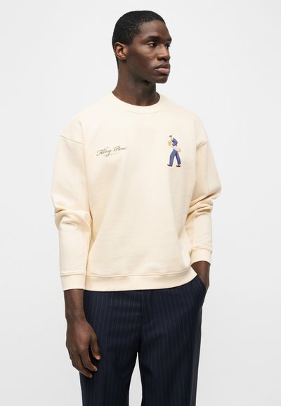 Άνδρας που φοράει εκρού φούτερ με μικρό γραφικό σχέδιο και κείμενο στο στήθος, σε συνδυασμό με μπλε παντελόνι με λεπτές ρίγες, στέκεται με το ένα χέρι στην τσέπη.