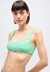 Νεαρή γυναίκα με κοντά καστανά μαλλιά φορά πράσινο αθλητικό σουτιέν υφής, στέκεται μπροστά σε απλό λευκό φόντο.