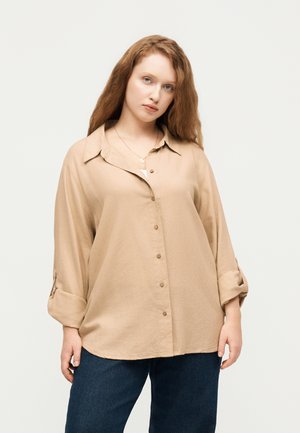 Junge Frau mit lockigem rotem Haar, die ein beiges Hemd mit Knöpfen und hochgekrempelten Ärmeln sowie eine dunkelblaue Hose trägt, vor hellem Hintergrund stehend.