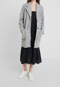 Harmaa teksturoitu pitkä neuletakki, jossa on käännetty kaulus, kaksi etutaskua ja yksinäppäin suljenta, päällä mustan pilkullisen mekon ja valkoisten lenkkareiden päällä.