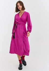 Długa, różowa sukienka kopertowa z marszczonymi rękawami, w groszki, z paskiem w talii, z rozcięciem po boku oraz gładką fakturą. Połączona z czarnymi butami i czerwonymi skarpetkami.