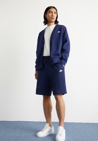Mornarsko modri cardigan z gumbi, v kombinaciji z usklajenimi kratkimi hlačami in belo majico. Na prsih in kratkih hlačah je embljem Nike. Bele športne copate.