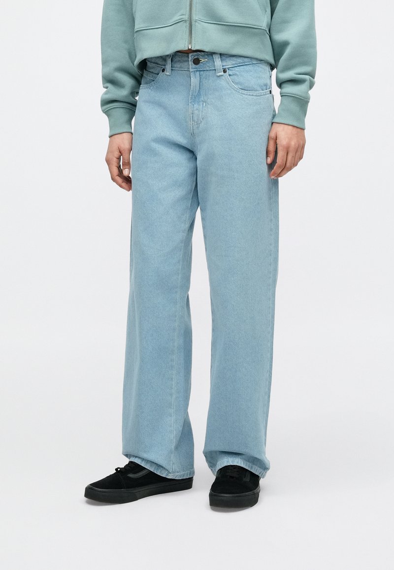 Persoon draagt lichtblauwe wijde jeans, zwarte sneakers en een groene zip-up trui, staand tegen een effen witte achtergrond.