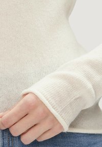 Mano che riposa vicino alla tasca dei jeans blu, indossando un maglione a coste di colore crema con maniche lunghe che si estendono oltre il polso.